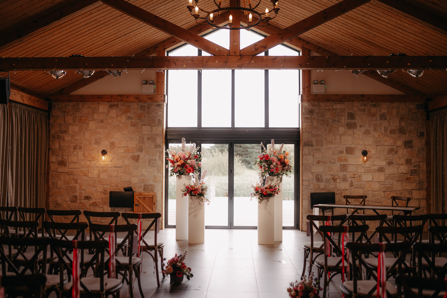 Barn wedding ceremony with stone walls, wooden beams, floral plinths and rows of chairs facing glass doors.